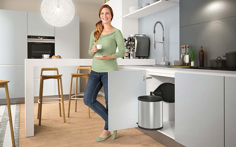 A woman is standing next to a waste bin in an open kitchen base cabinet