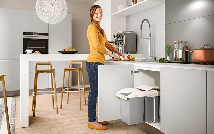 A woman is standing next to a built-in waste bin in an open kitchen base cabinet