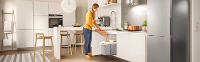 A woman stands in a kitchen in front of a built-in waste bin and throws vegetable waste into it.
