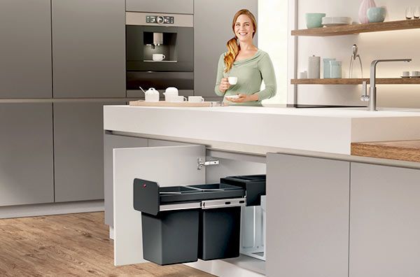 A woman is standing next to a waste bin in an open kitchen base cabinet