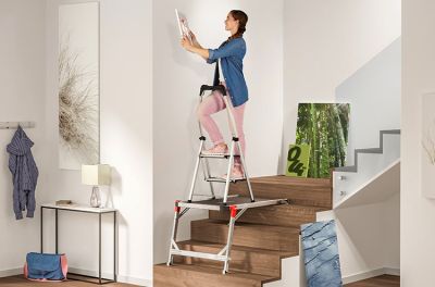 A person hangs a picture in the stairway while standing on a folding step that is placed on a stairs platform