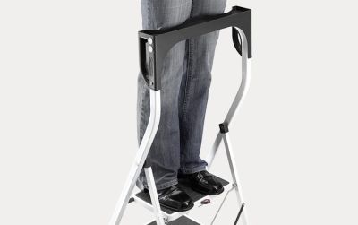 A woman is standing on the top step of a folding step and is leaning against the safety rail.
